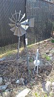 Windmill weathervane mounted outdoors in garden bed with rock mulch and fence behind.