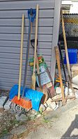 Full view of the entire collection of garden tools including shovels, brooms, pick axe, root feeder, and edger, arranged outdoors near a shed.