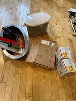 Lot overview showing metal tray with garden clippers, plastic storage bin, gardening themed cardboard box, and two boxed outdoor green floodlights on hardwood floor.
