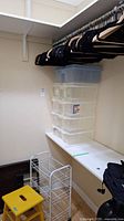 Wooden and black velvet hangers on a closet rod, clear plastic storage boxes stacked on counter, yellow foot stool on floor, and white metal wire drawer organizer next to stool.