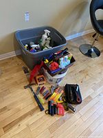 Photo showing a gray plastic tub with various cleaning sprays and liquid bottles. Adjacent white bucket on floor contains a brown toolbox and more cleaning supplies and hand tools.