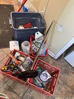 Photo showing various home maintenance items including a red-handled mop, large blue tote, red crates with assorted hardware and tools, a small shovel, and white grout float.