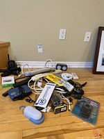 View of assorted household electronics on floor including flip phone, power strips, blue container, clock radio, barometer, chargers, and adapters.