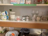Shelved display showing a variety of kitchenware including ceramic cups, egg coddlers, glass stemware and barware, and a cinnamon shaker.