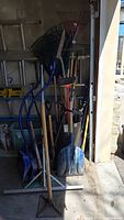 Photo showing multiple garden tools including shovels, rakes, broom, and tamper clustered in garage corner