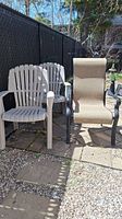 Three outdoor patio chairs: two beige plastic Adirondack-style chairs and one beige mesh fabric chair with dark metal armrests and frame on paved stone and gravel ground against a black fence.