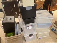 Stacked plastic storage bins in black, transparent, beige, and grey with larger and smaller sizes to the side and front, showing details of latching lids and labels on some boxes