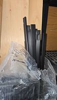 Close-up showing top ends of black PVC slats, flat with a folded tubular shape, inside a clear plastic bag with partial label showing.