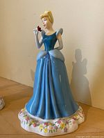 Front view of Cinderella figurine wearing blue gown holding red rose, showing facial detail and floral base.