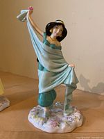 Front angle showing Jasmine holding light blue cloth with pink flower and standing on floral base.