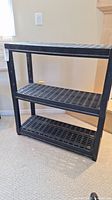 Front view of a black plastic shelf unit showing three perforated tiers and sturdy rectangular frame.