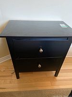 Front view of black wooden side table with two drawers and round metal knobs, showing the overall condition with scratches and scuffs.