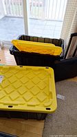 Three large black heavy duty plastic storage bins, with bright yellow lids placed on and beside the bins. The bins have reinforced ribbing and handles.