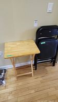 Wooden TV tray table next to stack of three black padded metal folding chairs against beige wall.