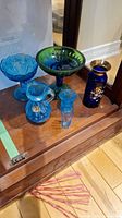 Set of six colored glass items including blue and green vases and compotes displayed on wooden surface.