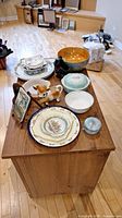 View of entire lot on wooden table showing vintage ceramic plates, bowls, decorative items including cow creamer and cat planter