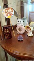 Photo showing white ceramic cat thermometer, vintage fan with girl illustration, ceramic little girl planter, brown lacquered jar, amethyst stone cluster.