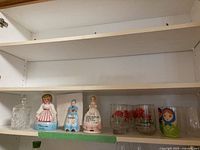 Shelf with vintage glass jar with lid, three figurative ceramic items including Kleanser Kate, ceramic dinner bell, ceramic woman figure, three carnation-themed glasses, and wooden Russian nesting doll.