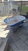 Rusty heavy duty metal wheelbarrow with wooden handles on paved ground with metal fence in background.