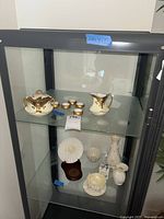 Photo showing upper shelf with porcelain lidded dish, small cups with gold accents, and cream pitcher with gold handle.
