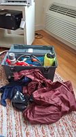 Plastic case on carpet filled with assorted camping gear including tools, rolled camping item, and red rain jacket nearby.
