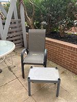 Photo showing one black metal framed patio chair with taupe mesh seat and back, set on a stone patio next to brick wall and lattice fencing. Small gray plastic side table in front of the chair, outdoor setting.