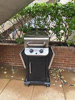Front view of a black Char-Broil gas grill with stainless steel lid and two control knobs, standing on patio with leafy plant background.