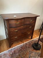 Wooden chest front view showing four drawers with brass handles, medium to dark stained wood, minor surface scratches, standing on wooden floor with a rug in front.