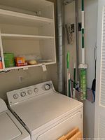 Wide view of laundry room showing white appliances, shelving, mops, broom, dust pan, sponges, and cleaning supplies