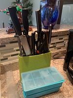 Green perforated plastic holder containing various knives, scissors, and peelers, next to two stacked blue ice cube trays with translucent lids on a kitchen counter.