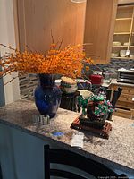 Blue glass vase holding tall orange faux plant arrangement with two clear glass candle holders in front and Lego plant on the side.