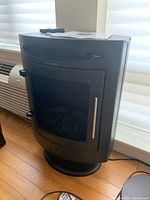 Front view of the black XLS Products electric fireplace heater standing on a wooden floor near a window, showing the closed mesh door and silver handle.