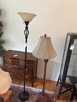 Photo showing two floor lamps side by side, one with a beige fabric shade and carved reddish wooden base, the other with a beige glass torchiere shade and black metal stand with decorative scrollwork on the stand.