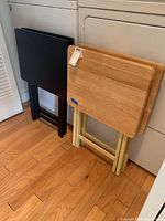 Four folding TV trays leaning against laundry machines, dark wood (black) and light wood with cream legs visible