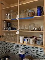 Two wooden shelves filled with various mugs, glass jars, glasses, and containers displayed in a kitchen cabinet.