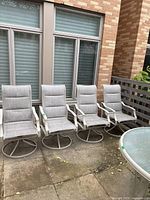Photo showing four matching outdoor patio chairs with cushioned seats and swivel metal bases arranged in a row on outdoor stone patio in front of windows.