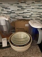 Wide shot of two plastic water filter pitchers, a set of mixing bowls stacked inside each other including two clear glass bowls, a white ceramic bowl, and a patterned brown ceramic bowl, with a box labeled ENID ramekins behind them on a kitchen counter with mosaic tile backsplash.
