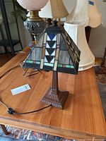 Side angle showing lamp shade's color and design pattern with geometric shapes and colors including white, black, green, red, and beige glass panels. Bronze finish base with square footed design visible.