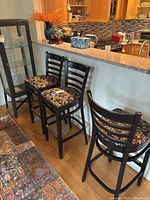 Three matching black wooden kitchen stools with ladder backs and floral cushioned seats placed near kitchen counter.