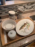 Photo of wood tray holding fish platter, canisters, nested white bowls, and woven placemats on a kitchen counter