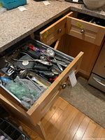 Open drawer filled with assorted kitchen utensils including spoons, peelers, and scissors, organized within metal utensil tray inside wooden kitchen drawer.