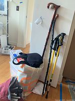 Walking sticks and hiking poles leaning against a wall, alongside a bag containing a black folding chair and other items