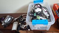 Plastic bin filled with assorted USB and charging cables, power adapters, and earbuds on a dark wood surface with some cables outside the bin.
