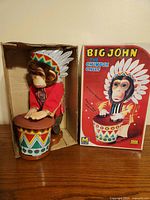 Photo showing the plush chimpanzee toy, dressed in Native American inspired clothing with feather headdress, playing the brightly decorated drum. The toy is positioned inside the original box with the box standing upright showing colorful graphics and title.