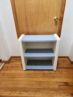 Small children’s bookcase with white sides and light blue shelves, plastic construction, standing against a wood door on hardwood floor