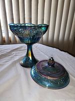 Blue carnival glass candy dish with lid off showing detailed grapevine and leaf pattern and scalloped edge.