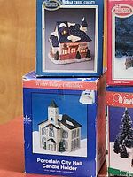 Four original boxes stacked, showing designs of light-up ceramic village buildings and accessories including Porcelain City Hall Candle Holder and a Station building