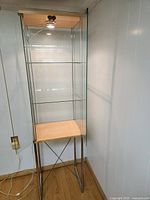 Full view of a tall, narrow glass display case. The case has three glass shelves and a wooden bottom shelf supported by silver metal legs. LED light is mounted on the top inside the case.