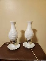 Pair of white milk glass hobnail textured lamps with ruffled edge bases on a brown surface against a beige wall.