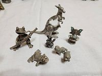 Photo showing seven metal animal and fairy figurines arranged on a white surface. Included are a kangaroo in mid-jump, a frog playing a banjo, a fairy with turquoise wings and pearl accent, two small dog figurines, and two small animal figurines.